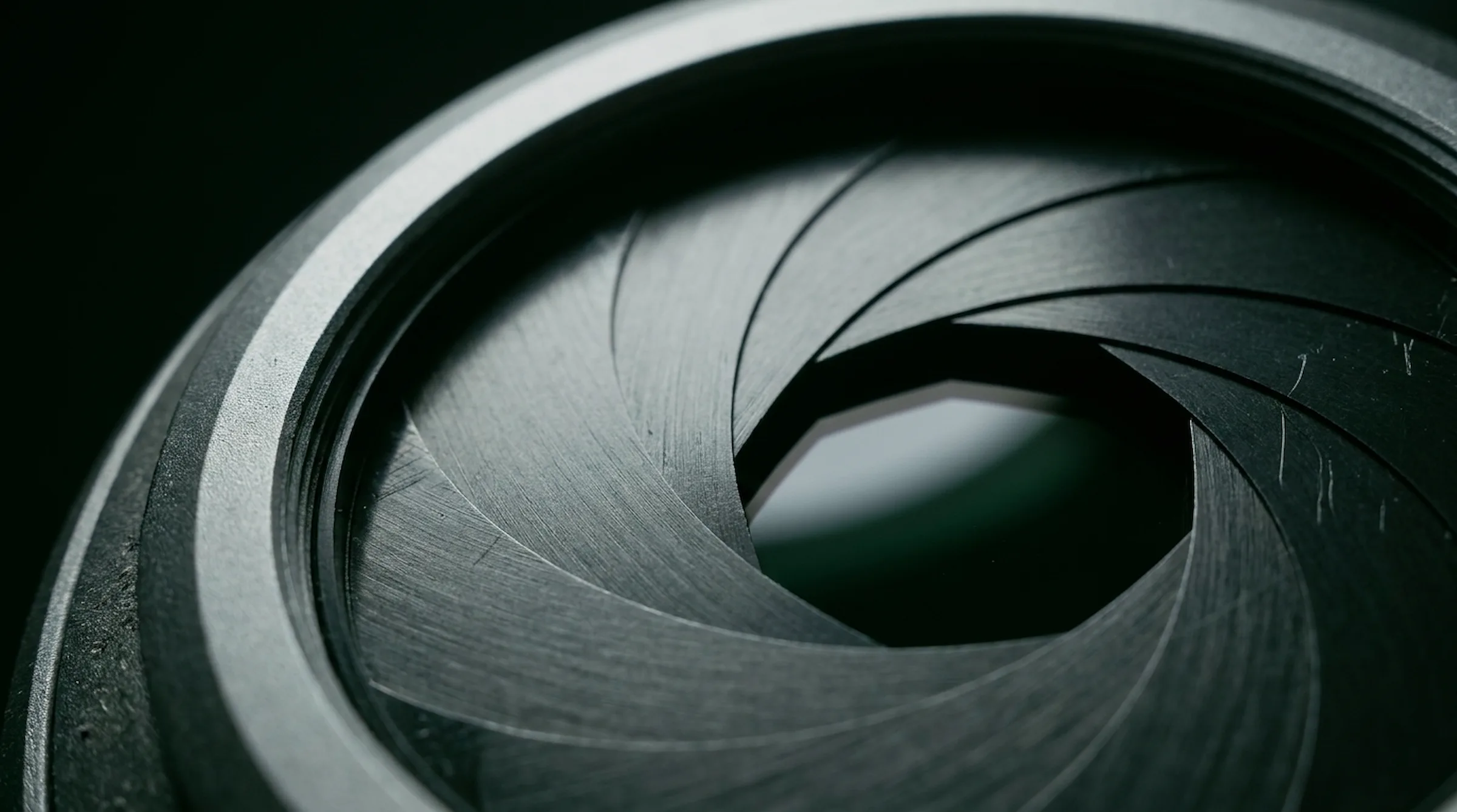 Macro render of camera aperture blades - dark machined metal with green-black reflections around a bright opening